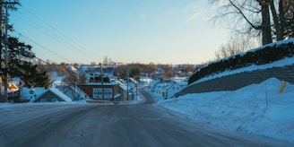 A snow-covered road leading into a small town during winter. The road is flanked by snowbanks and lined with houses. There is a signboard on the left for a general store. The sky is clear with a hint of the setting or rising sun.