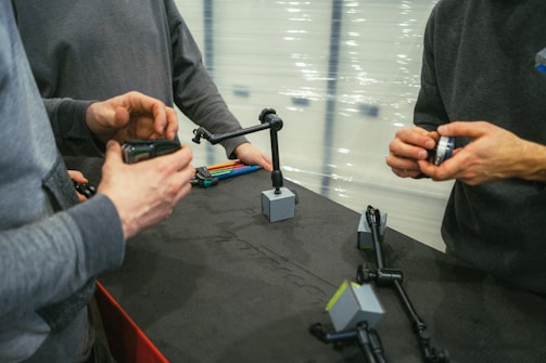 Three individuals are engaged in handling and assembling mechanical arms on a black surface. These extendable arms have base mounts in a cubic shape and appear to be part of a technical or engineering project. The individuals are focused and using their hands actively, implying collaboration or instruction.