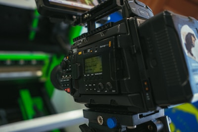 A professional video camera is set up with various controls and displays visible. The camera is positioned indoors with some green and metallic elements in the background, suggesting a setting like a studio or a set.
