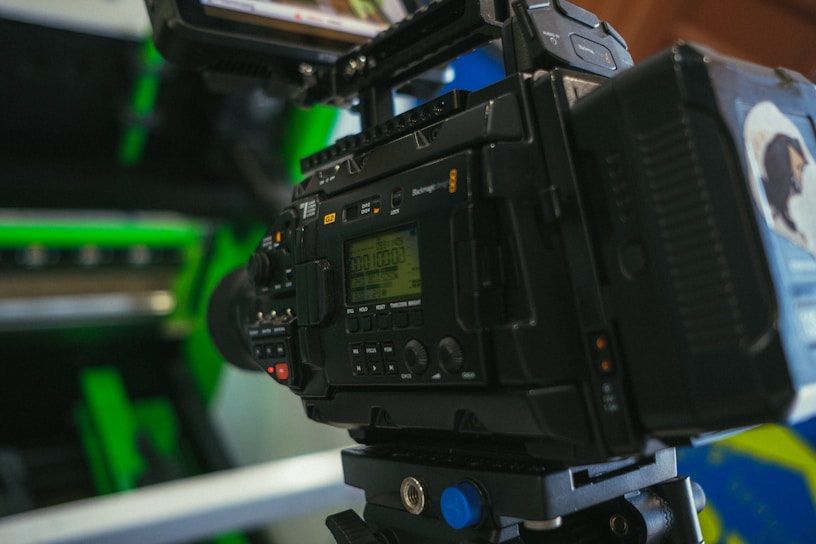 A professional video camera is set up with various controls and displays visible. The camera is positioned indoors with some green and metallic elements in the background, suggesting a setting like a studio or a set.
