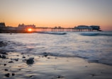 A scenic view of a seaside at sunset, featuring a long pier stretching into the calm sea. The sun is setting behind the pier, casting an orange and pink glow across the sky. Gentle waves lap at the rocky shoreline, creating a peaceful and serene atmosphere.