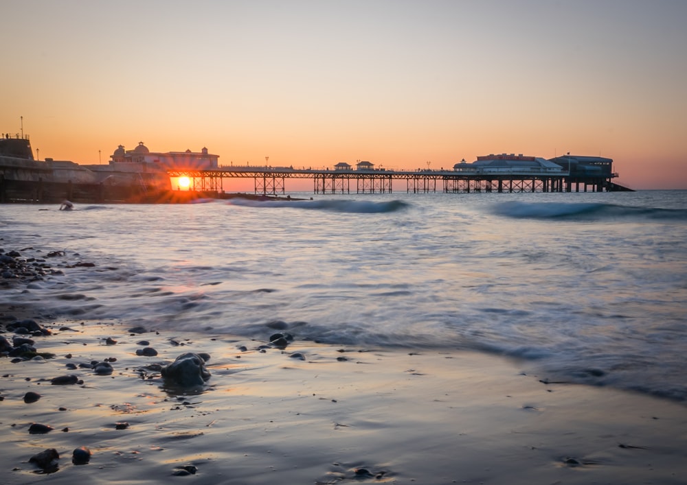 the sun is setting over the ocean with a pier in the background