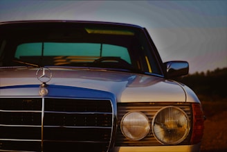 A vintage Mercedes-Benz car is captured in close-up, focusing on the front grille and headlights. The chrome details are polished and gleam under soft lighting, with a distinctive emblem prominently displayed in the center. The car is set in a dim, possibly evening environment with a subtle gradient in the sky, adding a classic and nostalgic aura.