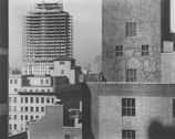 A view of urban architecture featuring several buildings. In the foreground, a building facade with visible cracks and patches can be seen, with four prominent windows. Behind it, there is a taller building under construction with a visible steel framework. The scene is devoid of people, highlighting the construction and architectural details.