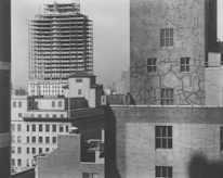 A view of urban architecture featuring several buildings. In the foreground, a building facade with visible cracks and patches can be seen, with four prominent windows. Behind it, there is a taller building under construction with a visible steel framework. The scene is devoid of people, highlighting the construction and architectural details.