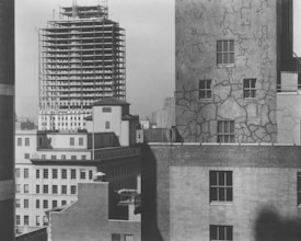 A view of urban architecture featuring several buildings. In the foreground, a building facade with visible cracks and patches can be seen, with four prominent windows. Behind it, there is a taller building under construction with a visible steel framework. The scene is devoid of people, highlighting the construction and architectural details.