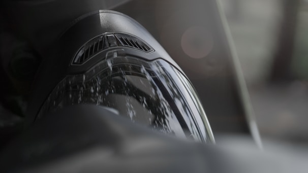 Close-up of a reflective motorcycle helmet visor with a soft focus background. The visor shows reflections of the surrounding greenery, while the top of the helmet includes a vented area.