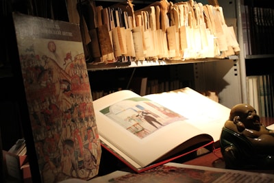 A dimly lit room with shelves filled with folders and books appears in the image. An open book, showcasing an illustration, rests on the table. A figurine, possibly a small statue of a laughing Buddha, adds character to the scene. The area has an academic or archival feel.