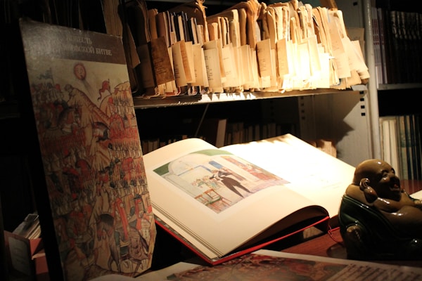 A dimly lit room with shelves filled with folders and books appears in the image. An open book, showcasing an illustration, rests on the table. A figurine, possibly a small statue of a laughing Buddha, adds character to the scene. The area has an academic or archival feel.