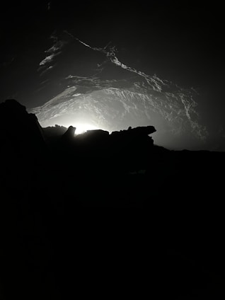 A dimly lit cave interior with rocky formations and a faint light source emanating from the background. Shadows and textures create an ominous and mysterious atmosphere.