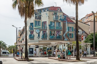 A vibrant mural covers the side of a building, depicting a lively scene with people on balconies engaged in various activities. The mural features cinematic elements, such as a film crew, cameras, and a person hanging from a clock, referencing classic movie moments. The building is located on a bustling street with palm trees and people walking and waiting at bus stops.