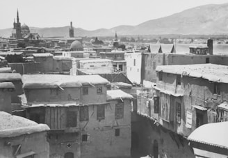 A historical cityscape with old, flat-roofed buildings made of stone or mud. Visible in the background are several minarets, domes, and a mountainous landscape, indicative of a Middle Eastern city. The architecture is traditional with streets winding between the buildings.