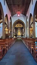 A grand and ornately decorated church interior featuring high vaulted ceilings and a long central aisle lined with wooden pews. The walls are adorned with intricate arches and columns. At the far end, an elaborate altar with religious icons and golden accents is illuminated with soft, ambient lighting.