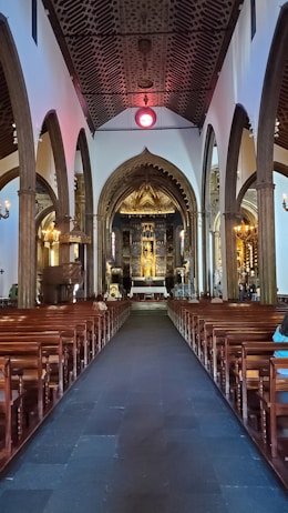 A grand and ornately decorated church interior featuring high vaulted ceilings and a long central aisle lined with wooden pews. The walls are adorned with intricate arches and columns. At the far end, an elaborate altar with religious icons and golden accents is illuminated with soft, ambient lighting.