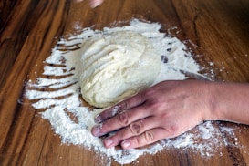 A hand is kneading dough on a wooden surface sprinkled with flour. The dough appears soft and is being shaped by the pressure of the hand.