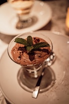 A decadent chocolate mousse served in a glass, garnished with fresh mint leaves and cocoa powder, placed on a white round plate. The lighting creates a warm and inviting atmosphere, with a blurred dessert in the background.