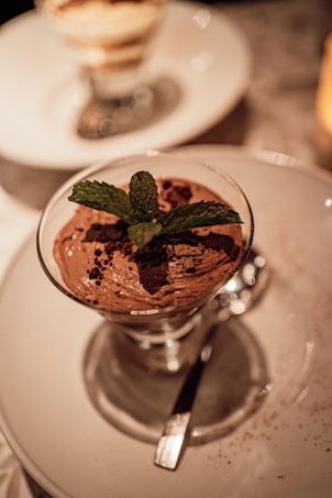 A decadent chocolate mousse served in a glass, garnished with fresh mint leaves and cocoa powder, placed on a white round plate. The lighting creates a warm and inviting atmosphere, with a blurred dessert in the background.