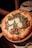 A freshly baked pizza topped with arugula, thin slices of cured meat, and large shavings of parmesan cheese. The crust is golden brown and slightly raised at the edges. The pizza sits on a wooden serving tray, and in the background, there is a blurred view of a table setting with white plates and cutlery.