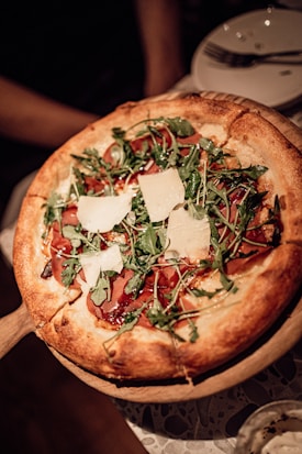 A freshly baked pizza topped with arugula, thin slices of cured meat, and large shavings of parmesan cheese. The crust is golden brown and slightly raised at the edges. The pizza sits on a wooden serving tray, and in the background, there is a blurred view of a table setting with white plates and cutlery.