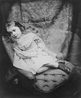 A young child with long hair is reclined on an upholstered chair. The child wears a checkered dress and lace-up boots. The expression conveys contemplation or introspection. The backdrop features a patterned fabric adding to the vintage atmosphere.