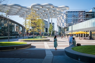 A modern urban plaza surrounded by contemporary architecture with curvaceous building designs. The plaza features circular grass patches, a unique canopy structure with cloud-like shapes above, and people casually walking or sitting in the area. Bright yellow umbrellas add a pop of color outside a café on the right.