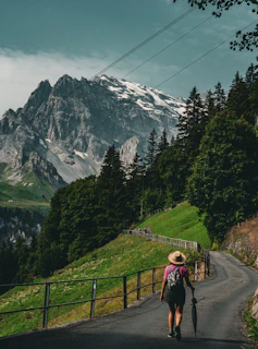 A person with a backpack and hat walks along a winding path surrounded by lush green hills and dense trees. Towering mountains with patches of snow rise in the background under a partly cloudy sky.