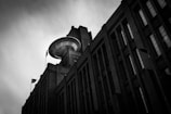 A dramatic black and white photograph of a large industrial building with an unusual circular structure on the roof, resembling a UFO. The building's tall, rectangular windows and brick facade are prominent in the composition, under a overcast sky. A flag is visible on one side of the building.