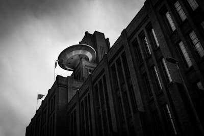 A dramatic black and white photograph of a large industrial building with an unusual circular structure on the roof, resembling a UFO. The building's tall, rectangular windows and brick facade are prominent in the composition, under a overcast sky. A flag is visible on one side of the building.
