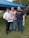 Three men standing together outdoors on a grassy area, with two of them holding certificates. They are under a blue canopy tent, and there are cars and trees in the background.