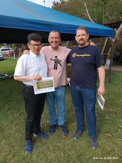Three men standing together outdoors on a grassy area, with two of them holding certificates. They are under a blue canopy tent, and there are cars and trees in the background.