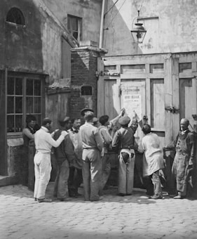 A group of men is gathered around a wooden gate, intently reading a large poster or notice that is affixed to it. The scene captures a moment of collective attention and engagement, with varied clothing indicating different professions or social classes. The setting is an outdoor area, bounded by a stone or brick structure, with a streetlamp suspended above.