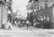 A black and white photograph capturing children playing in a cobblestone alley between two rows of wooden buildings. Several children are on a cart, while others engage in games on the ground. Laundry hangs overhead on lines strung between the buildings. The scene is lively, depicting a sense of community in an urban setting.