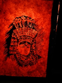 A highly detailed black ink illustration of a face adorned with an elaborate headdress, featuring multiple snake-like figures and intricate patterns. This artwork is set against a deep orange-red textured background that adds intensity to the image.
