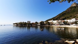 A serene coastal scene with calm waters reflecting the clear blue sky. A mountainous landscape with lush greenery rises to the right, while a small town with colorful buildings lines the shore. Boats are anchored near a small beach, adding to the tranquil atmosphere.
