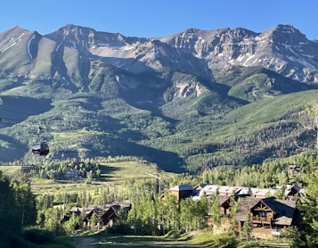 A picturesque mountain landscape featuring rugged peaks with patches of snow contrasted against lush green slopes. Below the mountains, a few rustic wooden cabins are nestled among dense green forests. A cable car system is visible, with a single car traveling along suspended cables, suggesting a ski resort or tourist area.