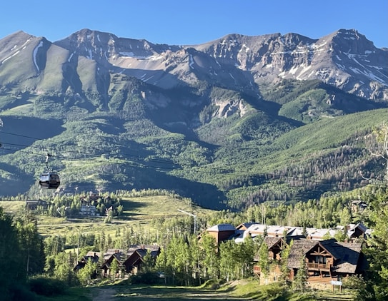 A picturesque mountain landscape featuring rugged peaks with patches of snow contrasted against lush green slopes. Below the mountains, a few rustic wooden cabins are nestled among dense green forests. A cable car system is visible, with a single car traveling along suspended cables, suggesting a ski resort or tourist area.