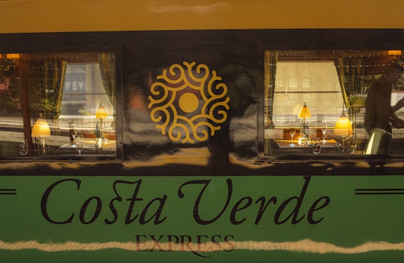 A train car with a luxurious interior is visible through two large windows. Inside, there are elegant table lamps, wine glasses, and dining setup. The exterior features the words 'Costa Verde Express' with a decorative emblem between the windows.