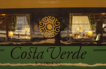 A train car with a luxurious interior is visible through two large windows. Inside, there are elegant table lamps, wine glasses, and dining setup. The exterior features the words 'Costa Verde Express' with a decorative emblem between the windows.