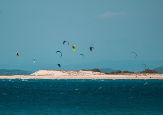 A group of colorful kite surfers gliding over the ocean, with an island or coastline in the background under a clear blue sky. The water is vibrant and deep blue, and there are several kiteboards visible.
