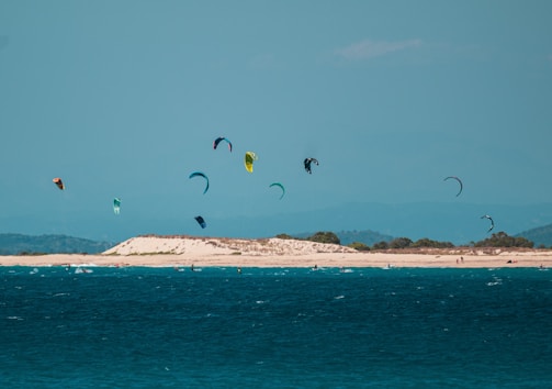 A group of colorful kite surfers gliding over the ocean, with an island or coastline in the background under a clear blue sky. The water is vibrant and deep blue, and there are several kiteboards visible.
