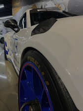 A close-up view of a sleek white sports car with blue rims and a Pirelli tire. The car has aerodynamic design elements with carbon accents and a visible side mirror. The background features a showroom setting with reflections on the polished floor and blurred images of other vehicles or display posters.