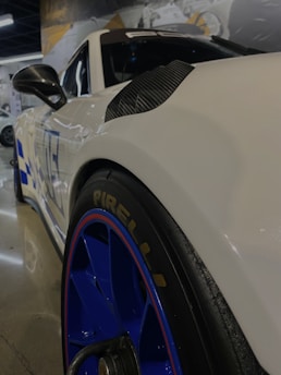 A close-up view of a sleek white sports car with blue rims and a Pirelli tire. The car has aerodynamic design elements with carbon accents and a visible side mirror. The background features a showroom setting with reflections on the polished floor and blurred images of other vehicles or display posters.