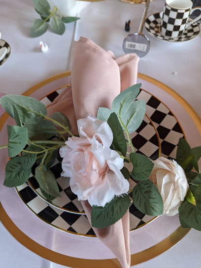 A decorative table setting features a folded blush napkin adorned with two artificial white roses. The arrangement rests on a checkered black and white plate, bordered by a gold rim. Surrounding objects include leafy green branches and additional tableware in a matching checkered pattern.