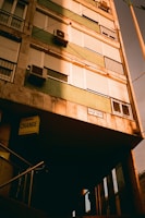A building facade with multiple windows, many of which have closed shutters. The building displays a mix of stone and green panels. Several air conditioning units are attached below the windows. A sign below indicates a currency exchange service and a street sign with the name 'József Körút' is visible. A set of metal railings leads to a shaded entrance.