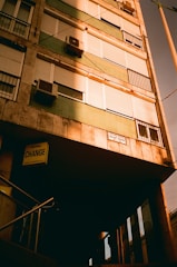A building facade with multiple windows, many of which have closed shutters. The building displays a mix of stone and green panels. Several air conditioning units are attached below the windows. A sign below indicates a currency exchange service and a street sign with the name 'József Körút' is visible. A set of metal railings leads to a shaded entrance.