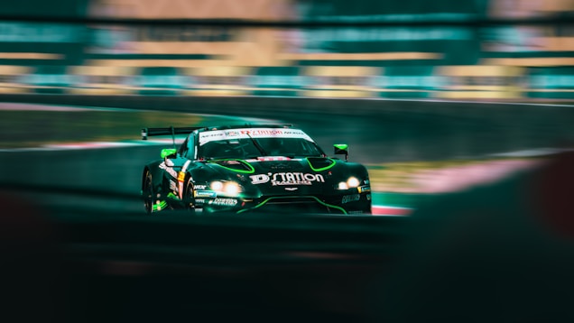 A sleek, high-performance racing car is speeding around a track. The car is predominantly black with green and neon accents, featuring various logos and sponsorship decals. Motion blur emphasizes its speed, while the background track and barriers add to the dynamic scene.