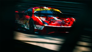 A sleek red sports car with branding and sponsor logos, speeding on a race track. The motion blur emphasizes its speed and dynamic motion, while the shadows cast by surrounding barriers suggest a sunny day at the race.