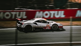 A sleek race car is speeding down a racetrack, with a blurred background that suggests motion. The car is predominantly white with red and black accents and features the number 54 prominently on its side. Advertising and branding are visible, with large logos and designs.