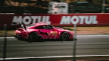 A sleek, high-performance sports car, painted in vibrant pink, speeds along a racetrack. Blurred motion lines in the background emphasize the car's speed. Advertising banners and a fenced-off area are visible along the track.