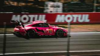A sleek, high-performance sports car, painted in vibrant pink, speeds along a racetrack. Blurred motion lines in the background emphasize the car's speed. Advertising banners and a fenced-off area are visible along the track.